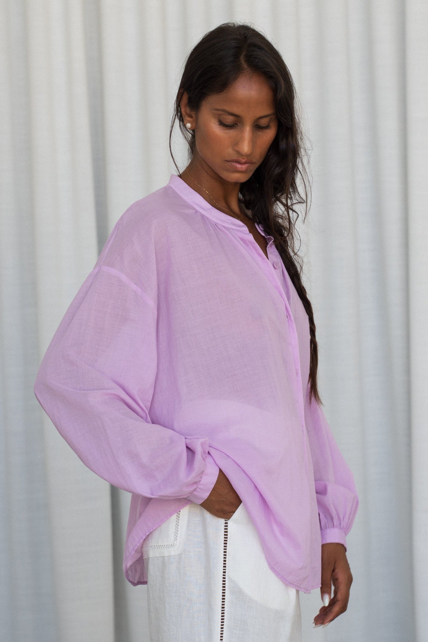 LILLY PILLY woman wearing a orchid Ember cotton voile shirt and white pants against a white curtain background