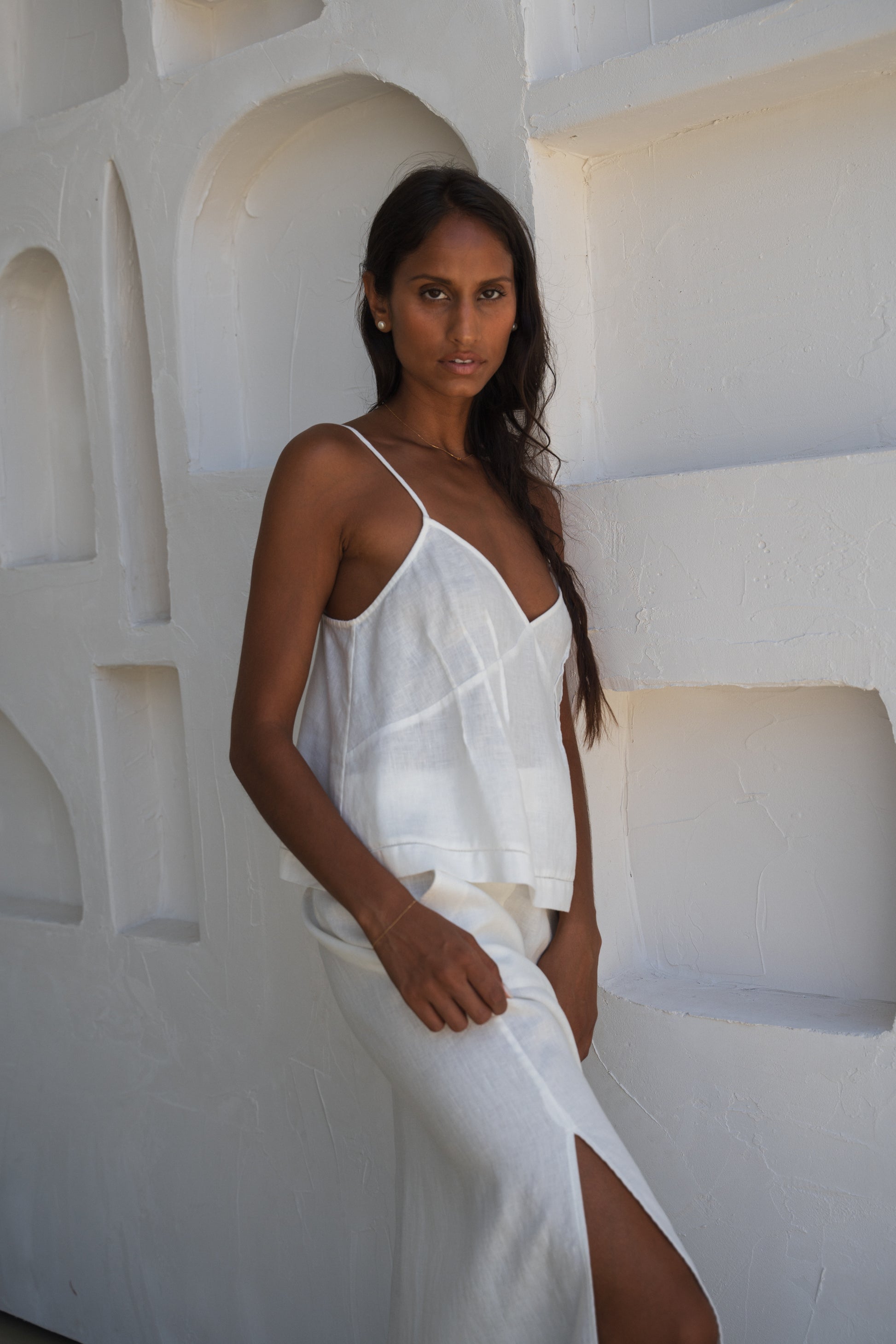 LILLY PILLY woman wearing a ivory Bea linen cami top and ivory skirt outfit standing against a textured white wall.