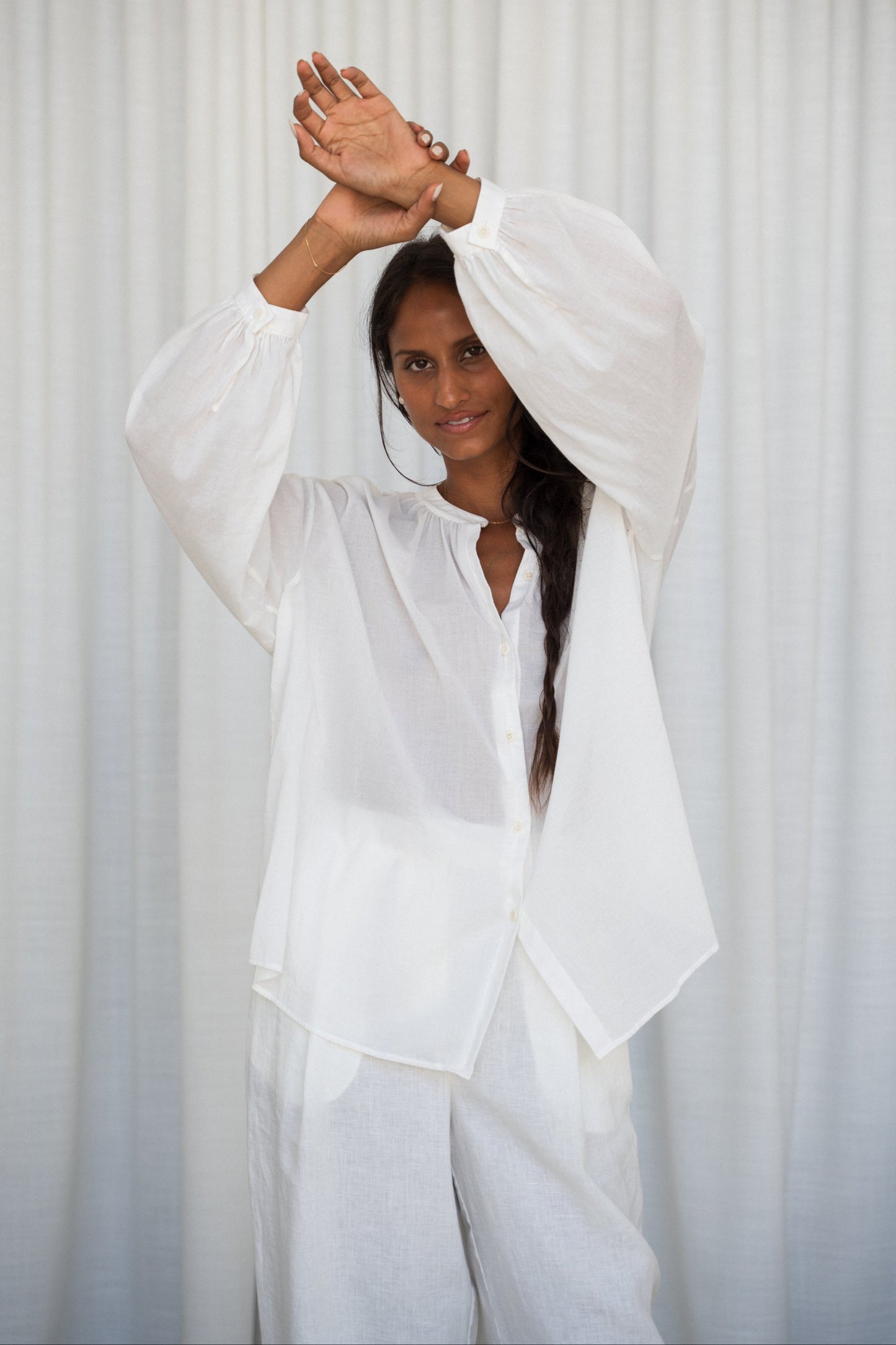 LILLY PILLY woman wearing a ivory Ember cotton voile shirt and ivory linen pants against a light curtain background