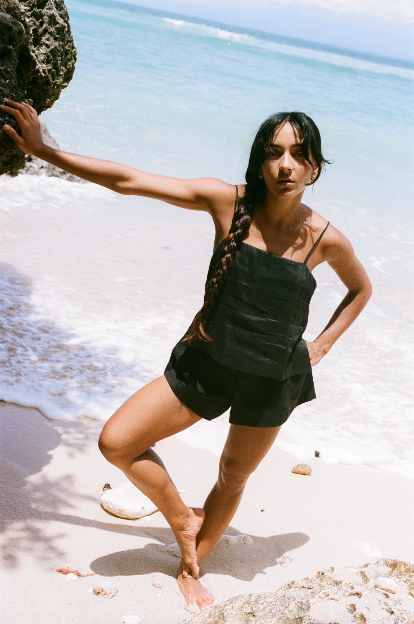 LILLY PILLY woman in black silk linen top and Gaia linen shorts standing on a beach with ocean waves in the background