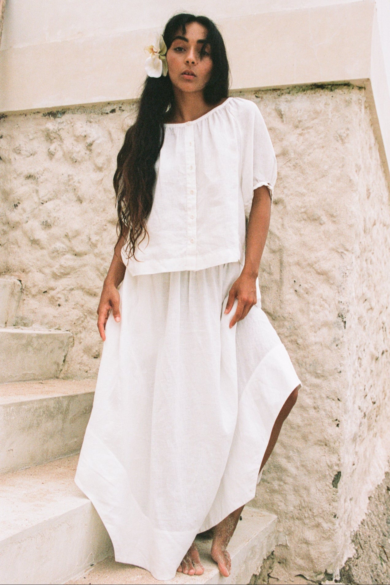 LILLY PILLY woman in an ivory Hannah linen skirt and Pia linen top standing on stone steps with a white flower in her hair.