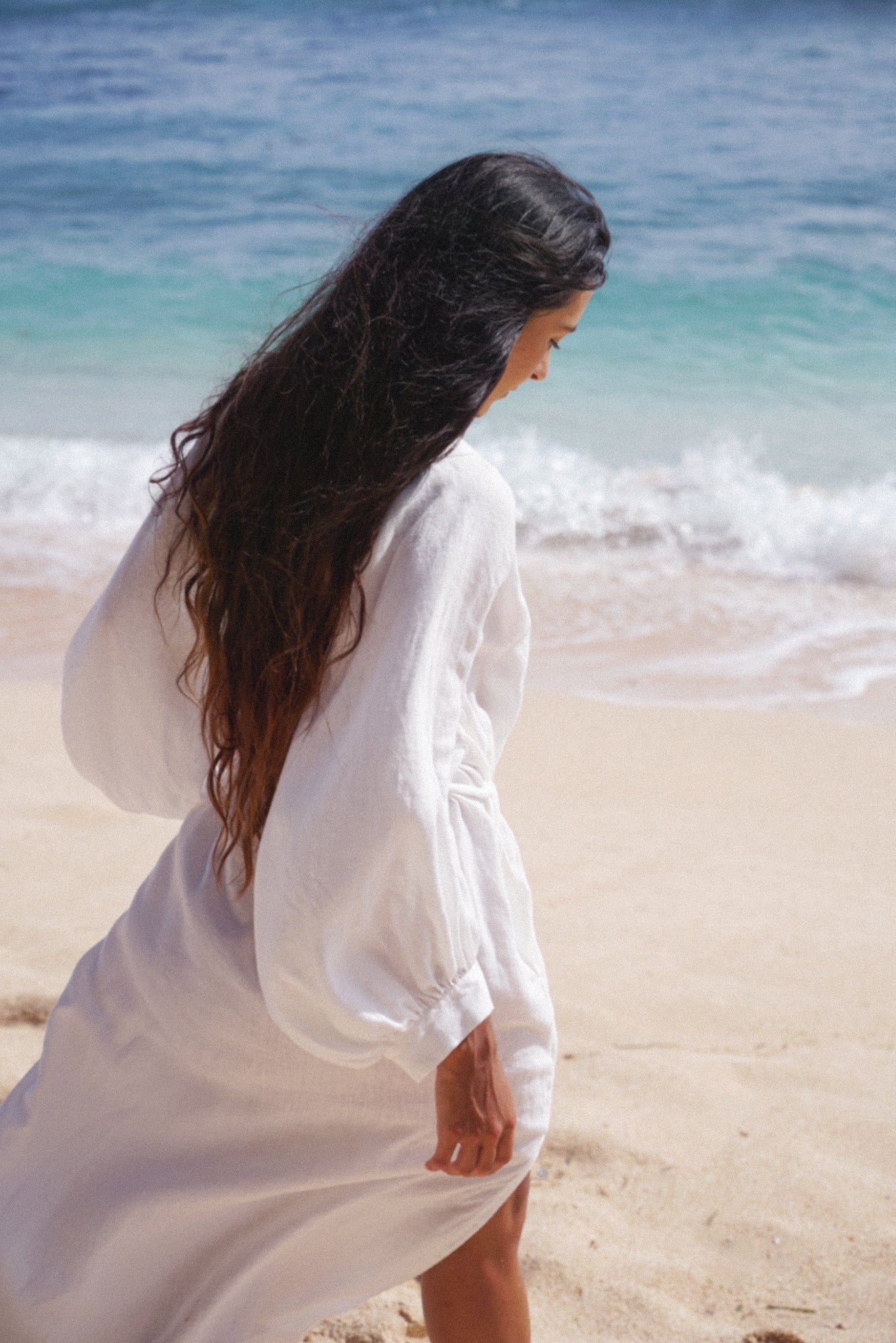 Woman in an ivory Mae linen dress standing on a beach with ocean waves in the background