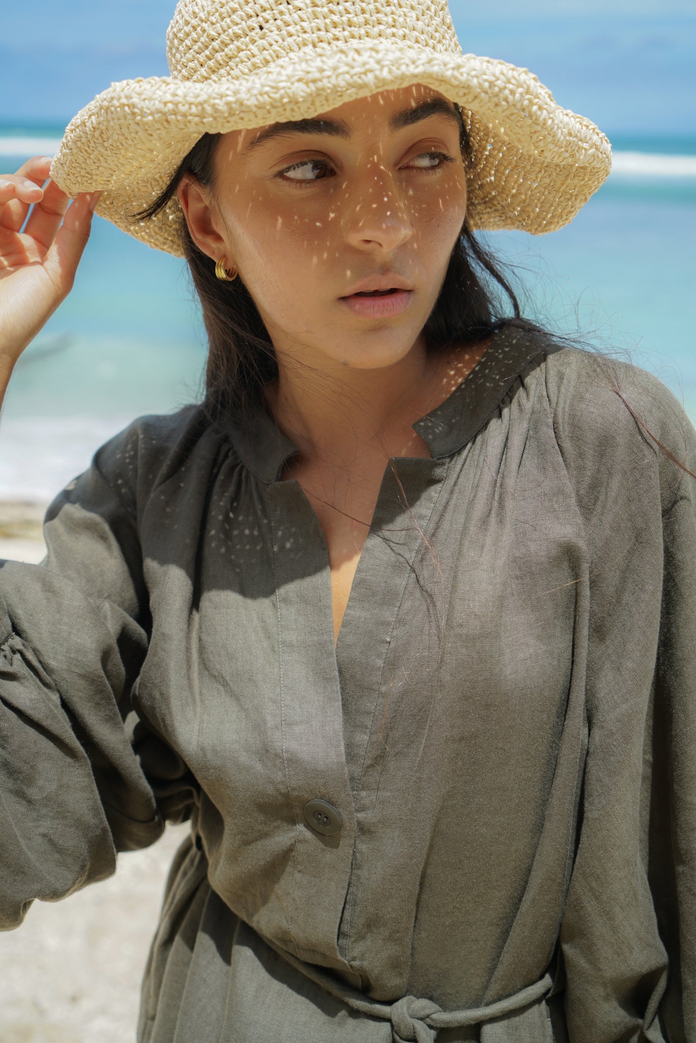 Woman wearing a khaki Mae linen dress on a beach with hat close up
