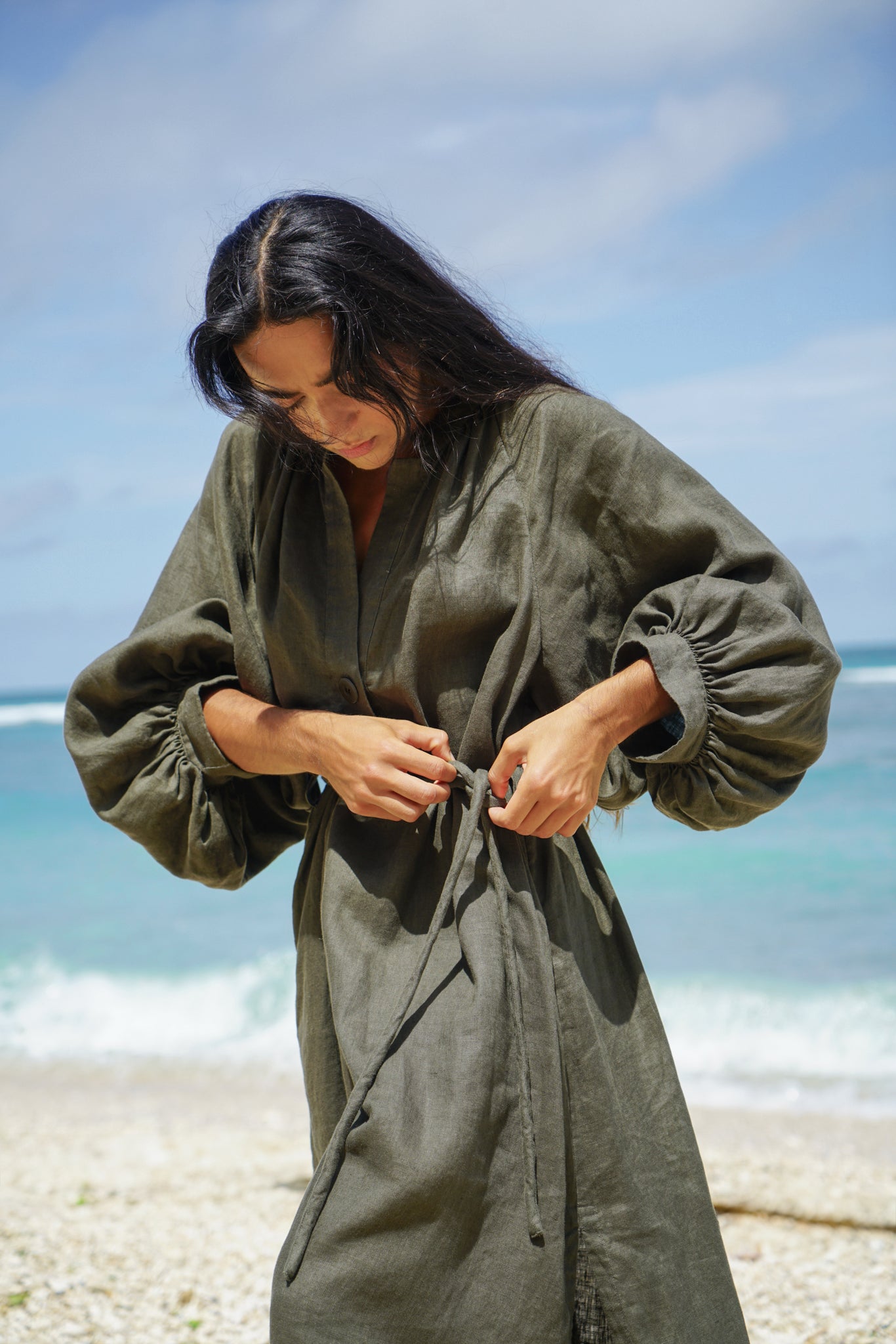 Woman wearing a khaki Mae linen dress on a beach with ocean and sky in the background