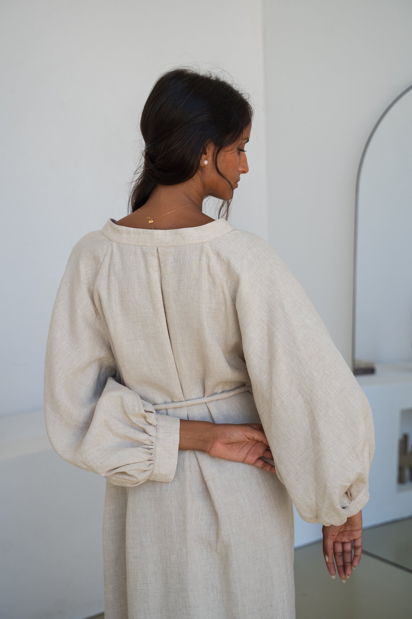 Woman wearing a Mae oatmeal linen dress close up back view in a room with white walls and a mirror.