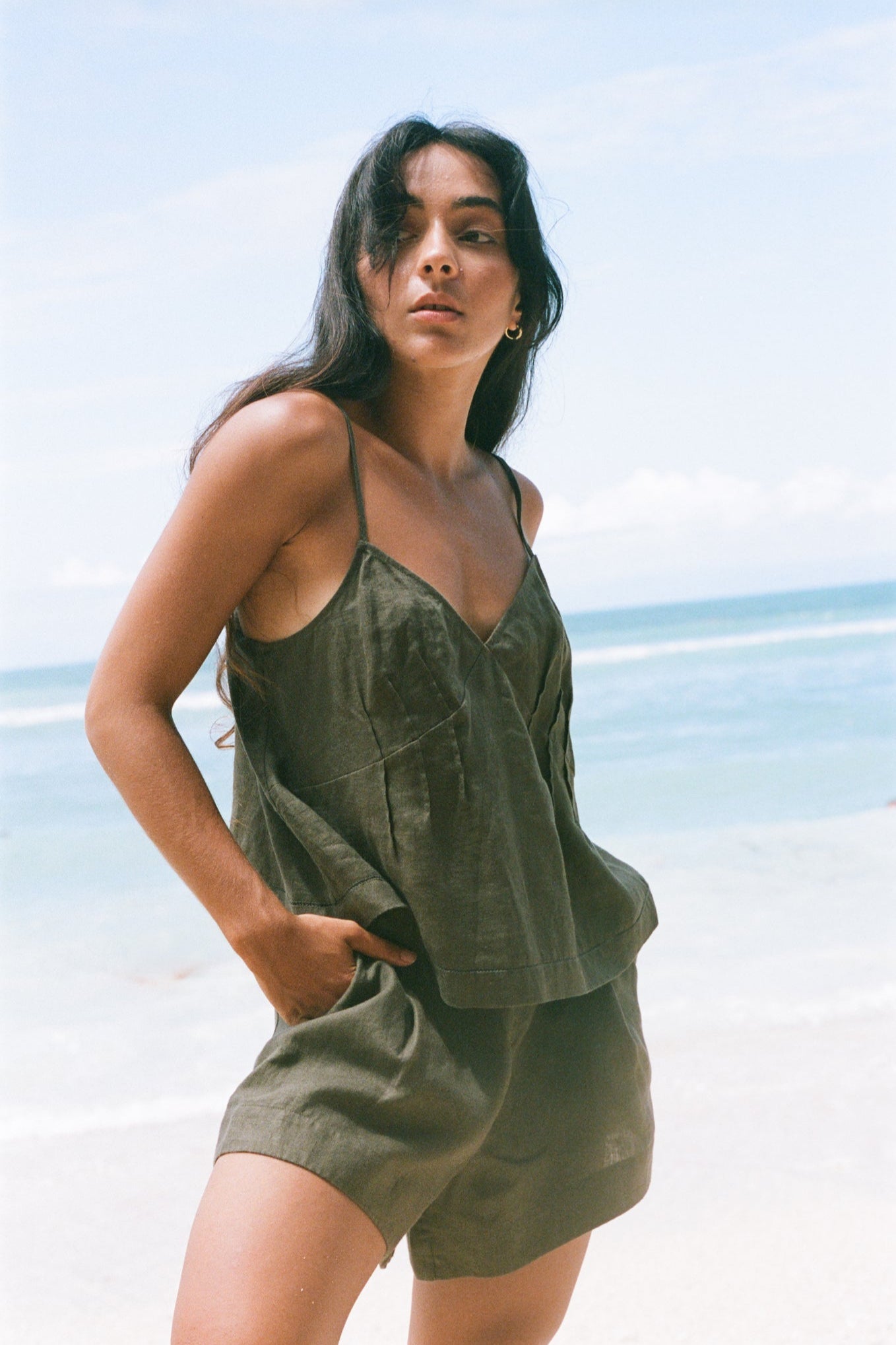LILLY PILLY woman wearing a khaki Bea linen cami top and Gaia shorts outfit on a beach with clear blue sky.
