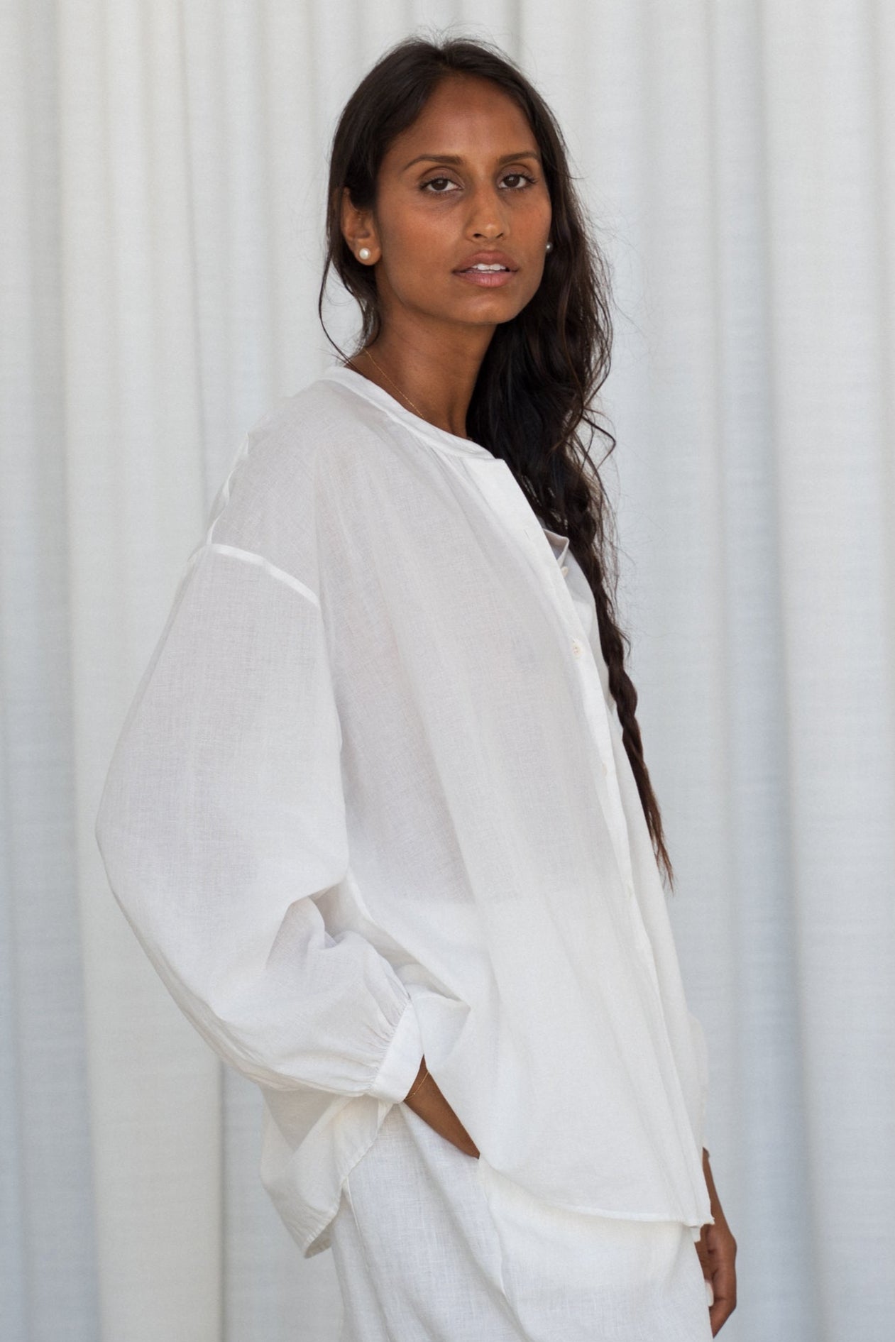 Close up of LILLY PILLY woman wearing a ivory Ember cotton voile shirt and ivory linen pants against a light curtain background