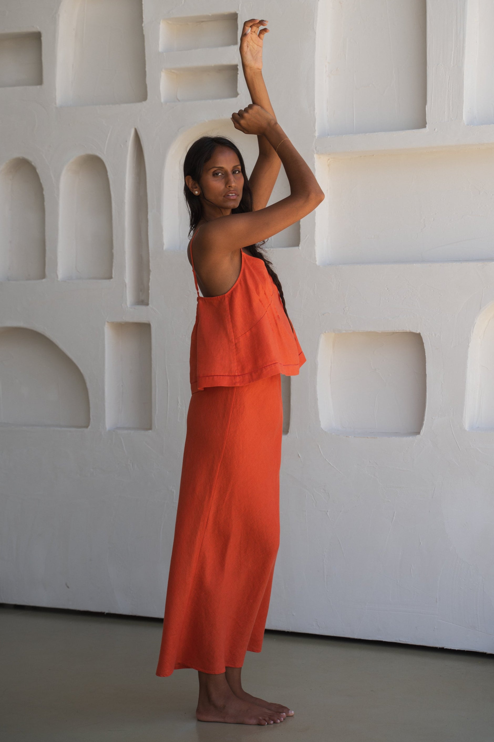 Close up of LILLY PILLY woman wearing a spritz Bea linen cami top with thin straps showing side view while posing against a textured white wall.