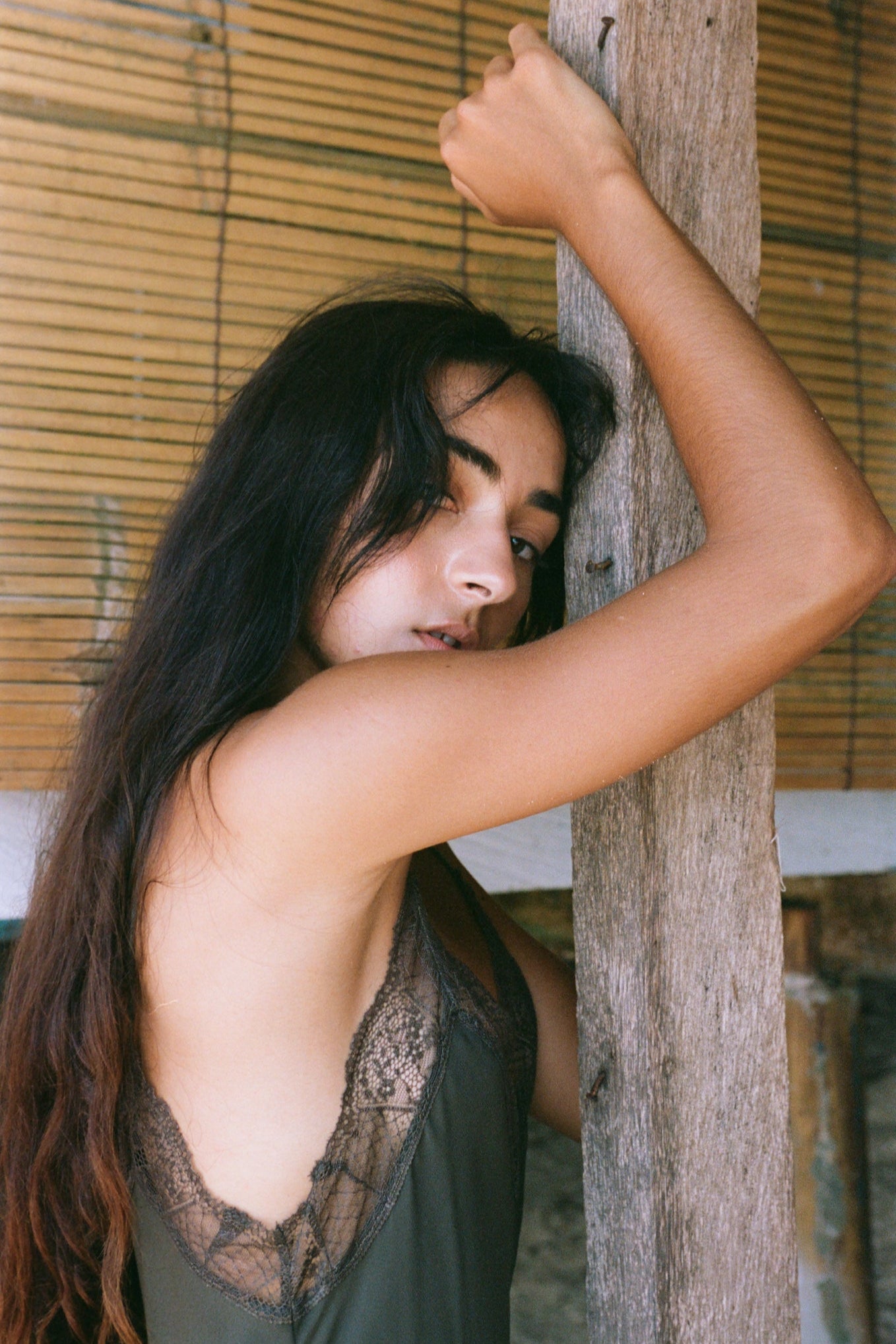 Woman in a khaki Freya silk slip dress leaning against a wooden post with bamboo blinds in the background