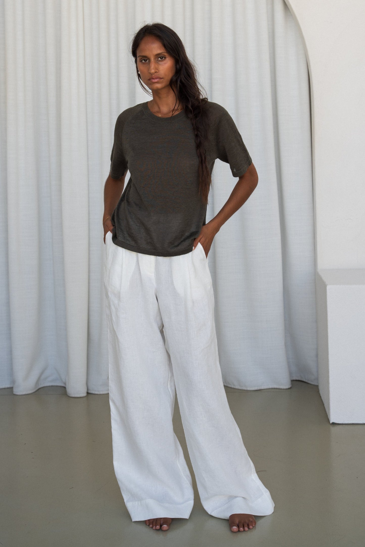 Woman wearing a khaki Grace linen knit top and Oli ivory linen pants with white curtain wall background