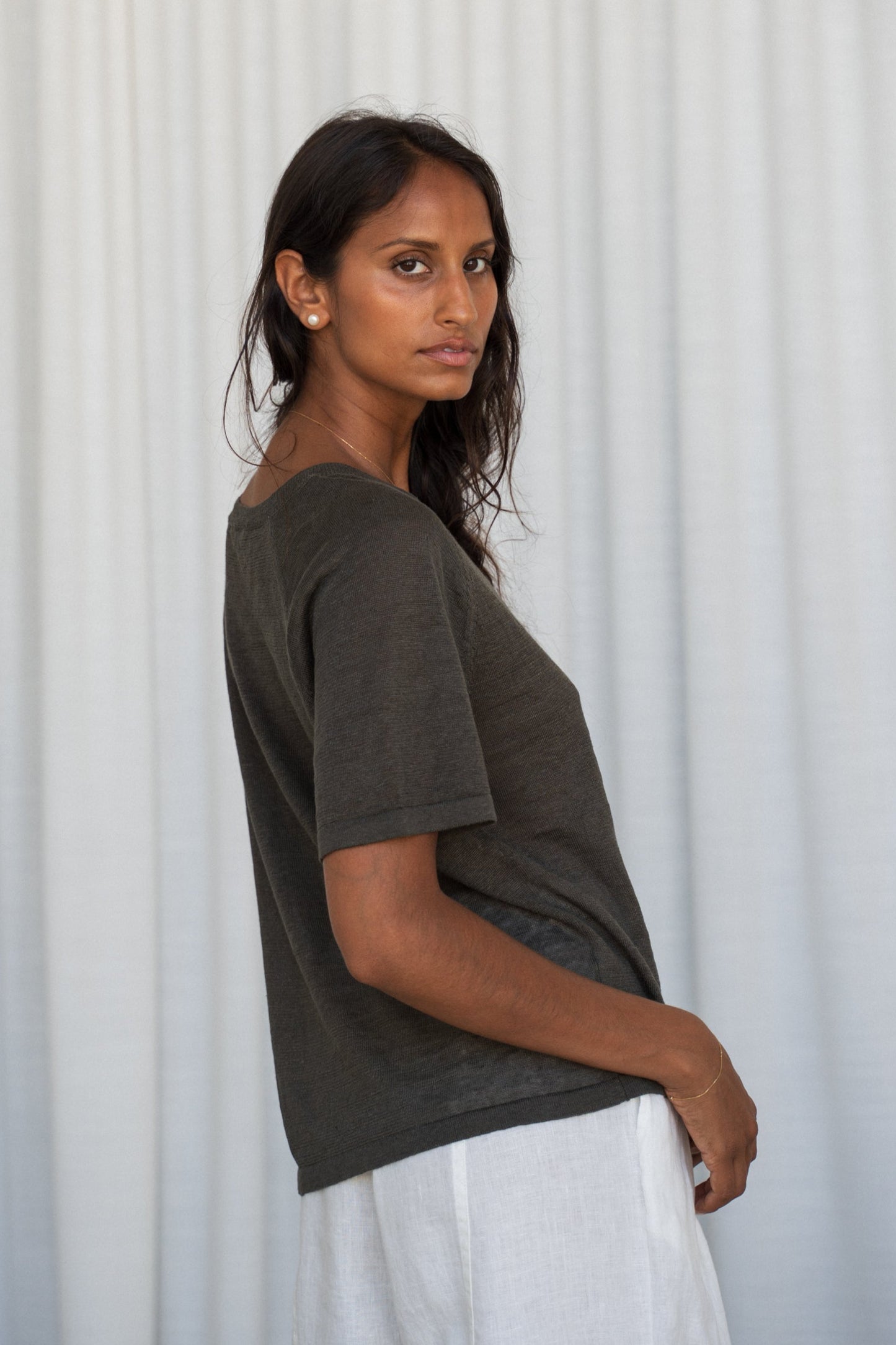 Woman wearing a khaki Grace linen knit top and Oli ivory linen pants, side view, with white curtain wall background