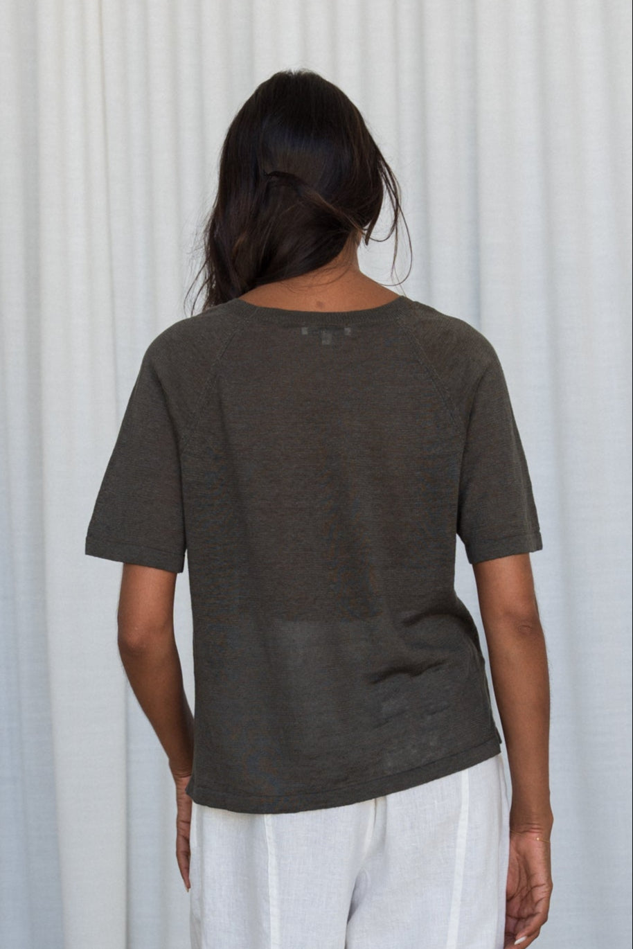 Woman wearing a khaki Grace linen knit top and Oli ivory linen pants back view with white curtain wall background