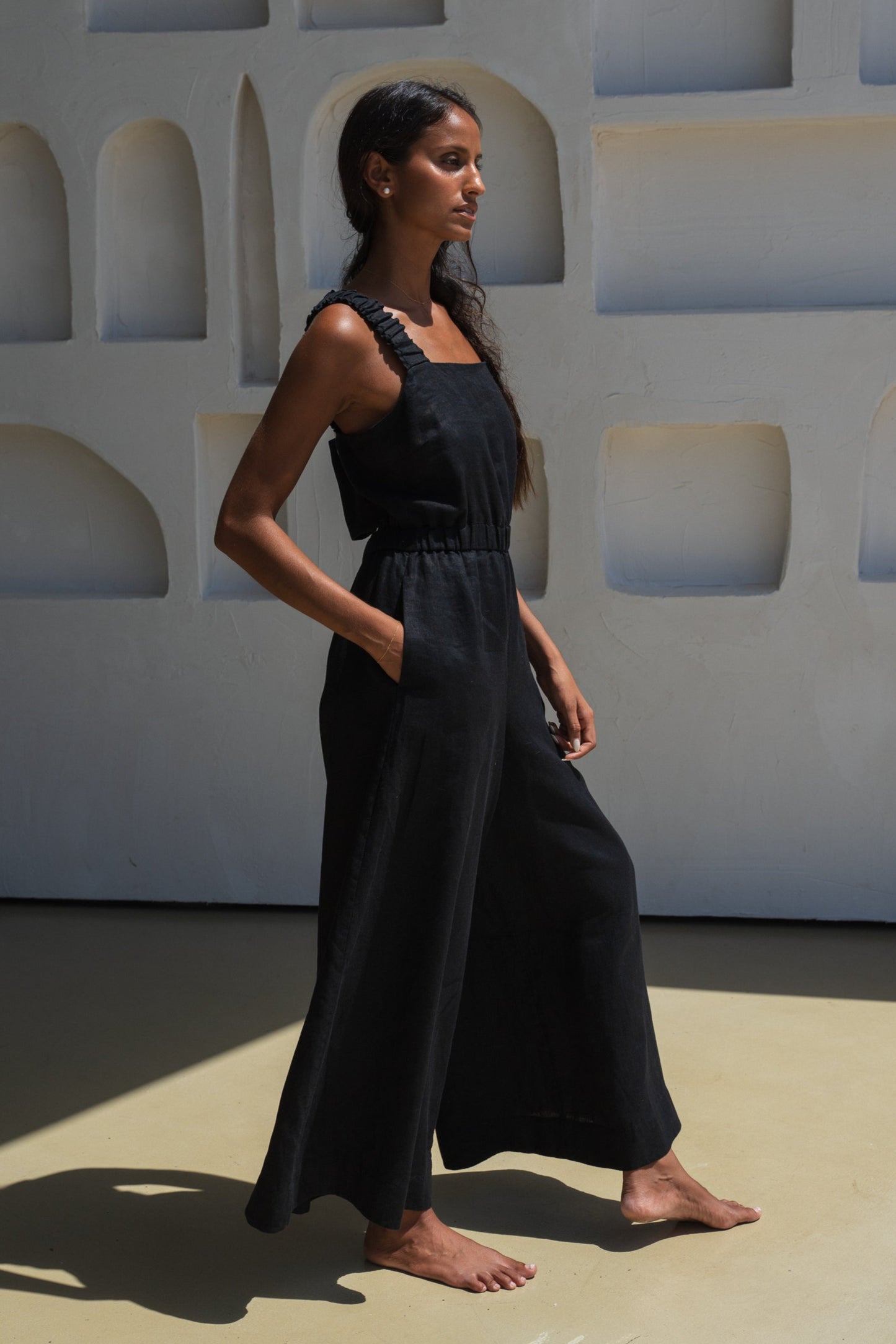 LILLY PILLY woman wearing a black Keely linen jumpsuit showing side view while standing against a geometric wall.