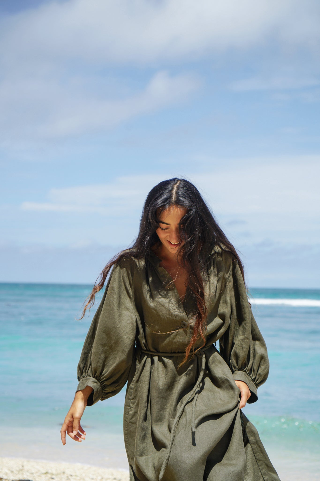 Woman wearing a khaki Mae linen dress on a beach with ocean and sky in the background
