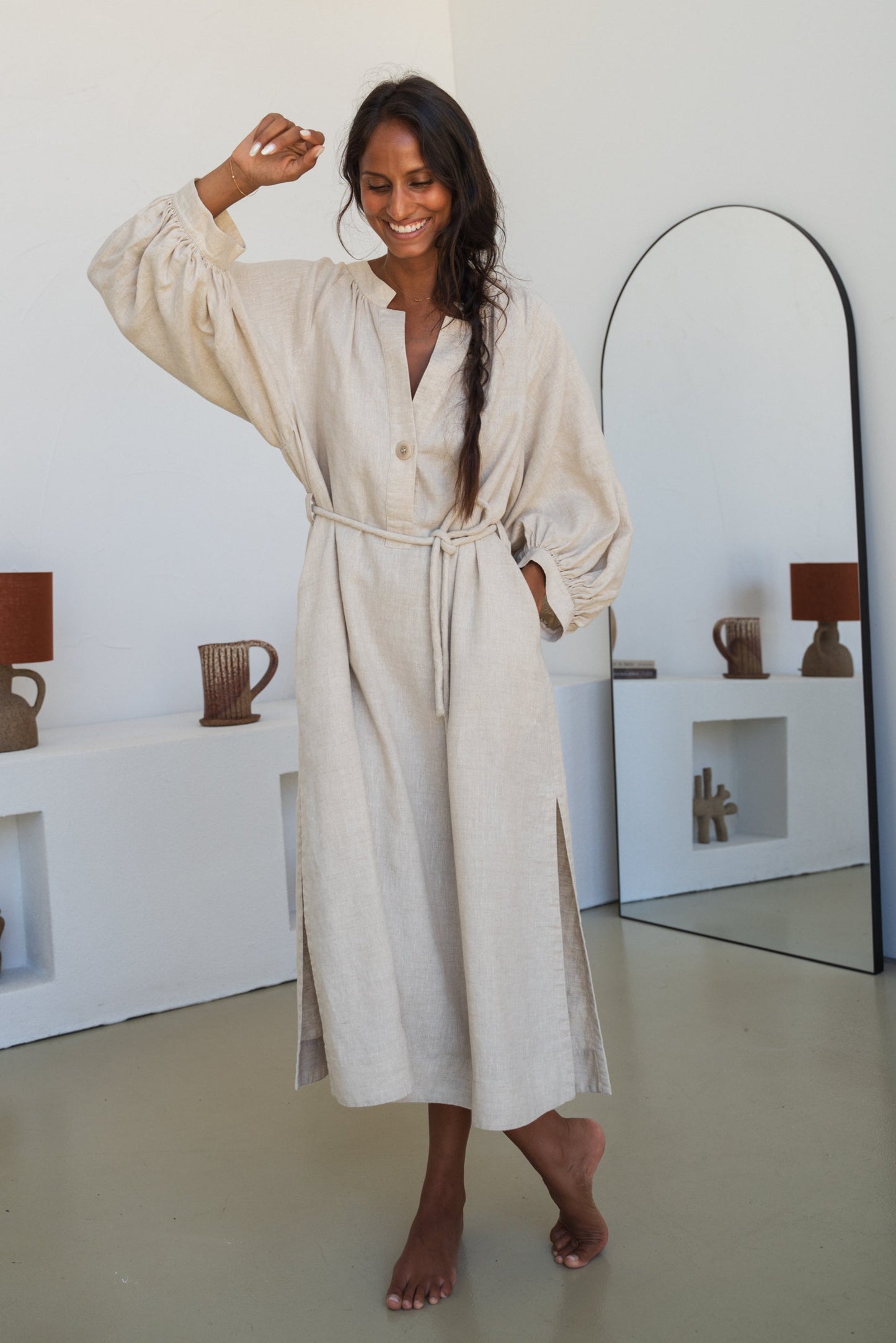 Woman wearing a Mae oatmeal linen dress standing in a room with white walls and a mirror.