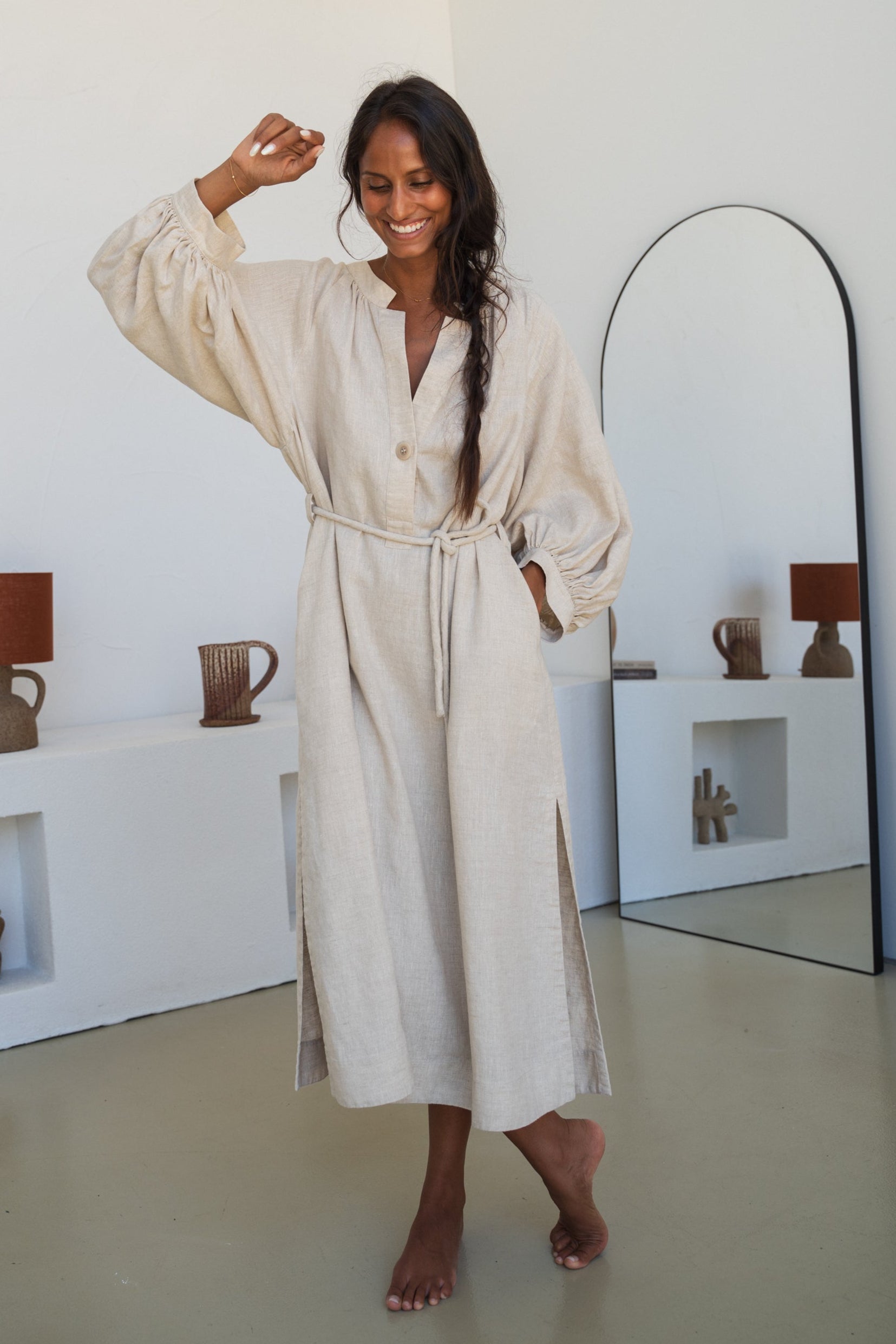 Woman wearing a Mae oatmeal linen dress standing in a room with white walls and a mirror.