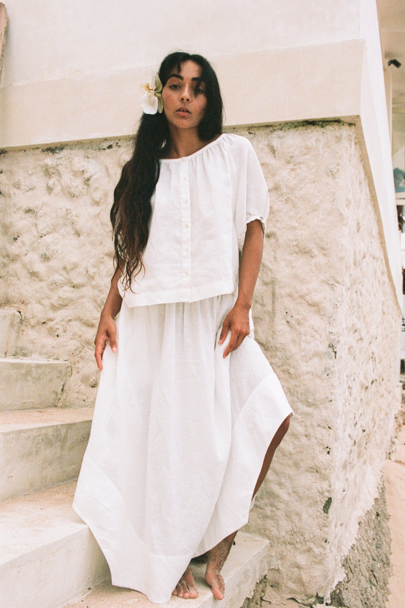 LILLY PILLY woman wearing an ivory Pia linen shirt top with ivory skirt standing on stone steps with a white wall in the background