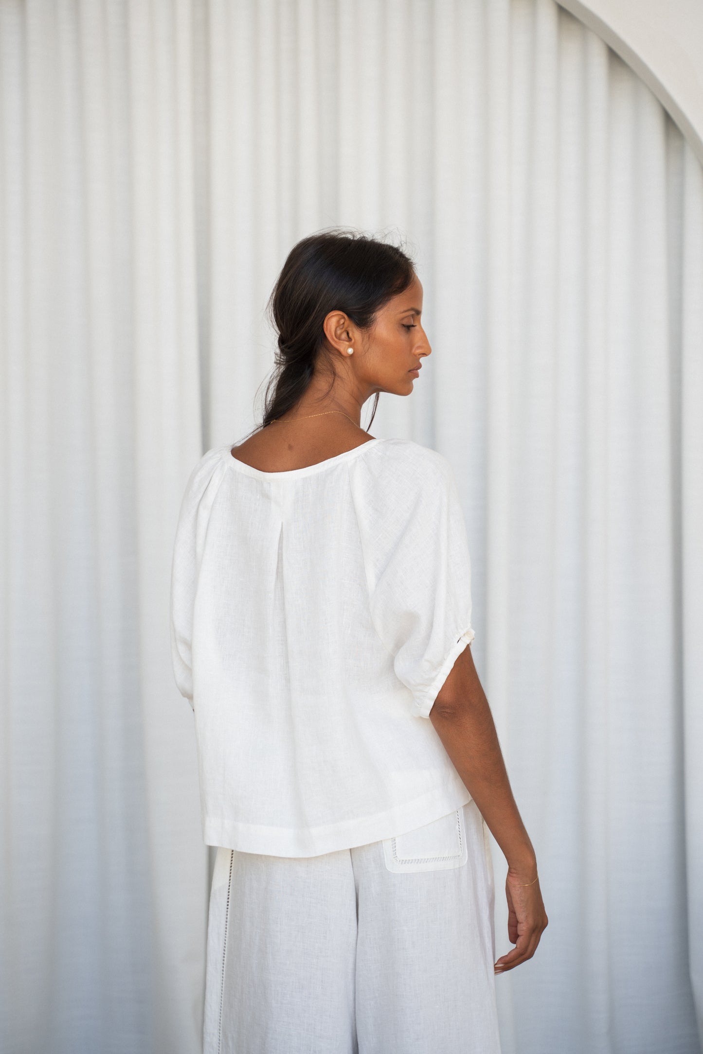 LILLY PILLY woman wearing an ivory Pia linen shirt top with ivory pants back view against a white curtain background