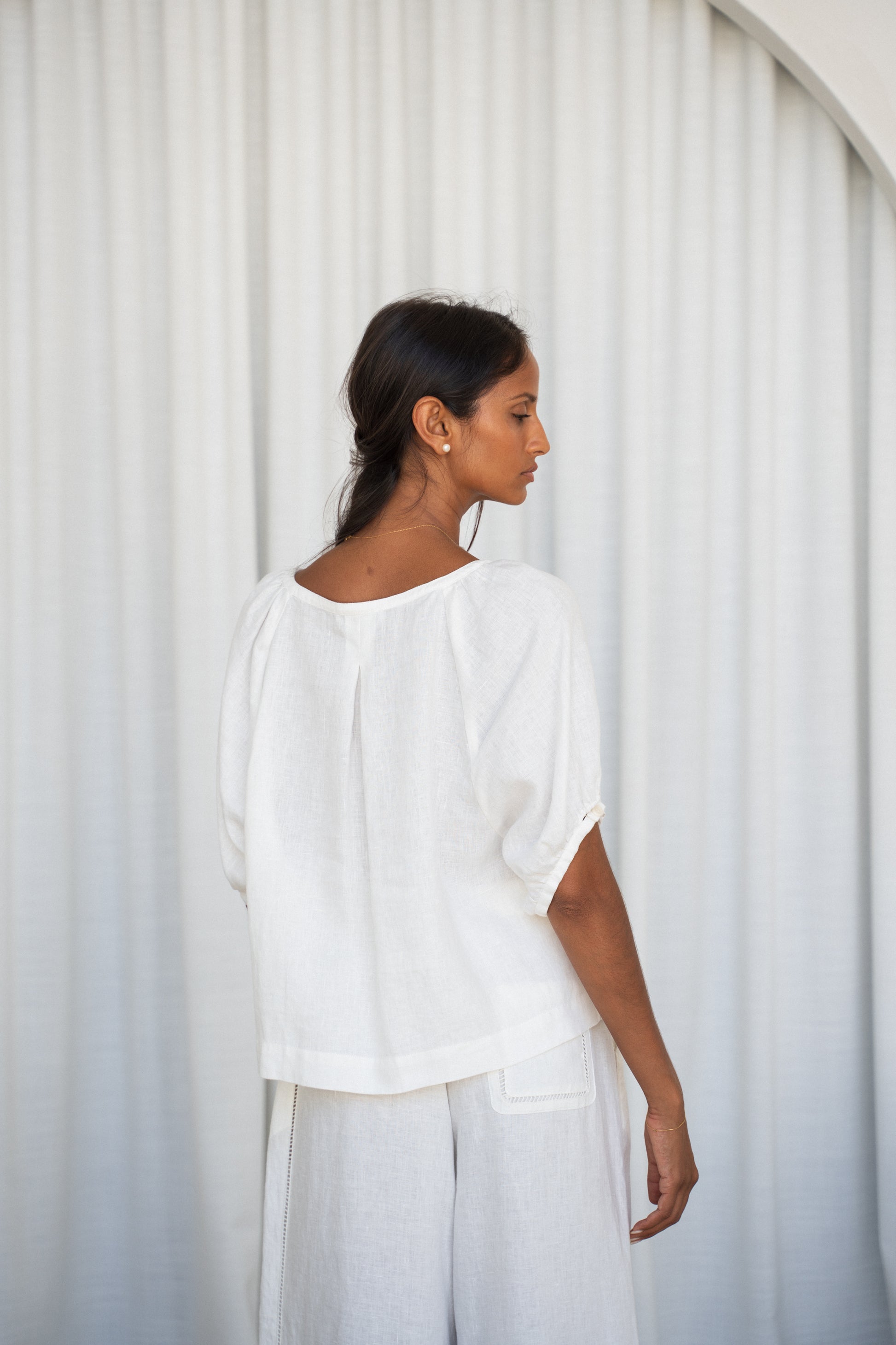 LILLY PILLY woman wearing an ivory Pia linen shirt top with ivory pants back view against a white curtain background