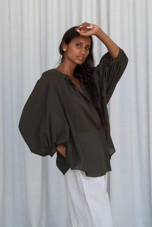 LILLY PILLY woman wearing a khaki Thalia cotton voile shirt blouse showing side view in front of a curtain background