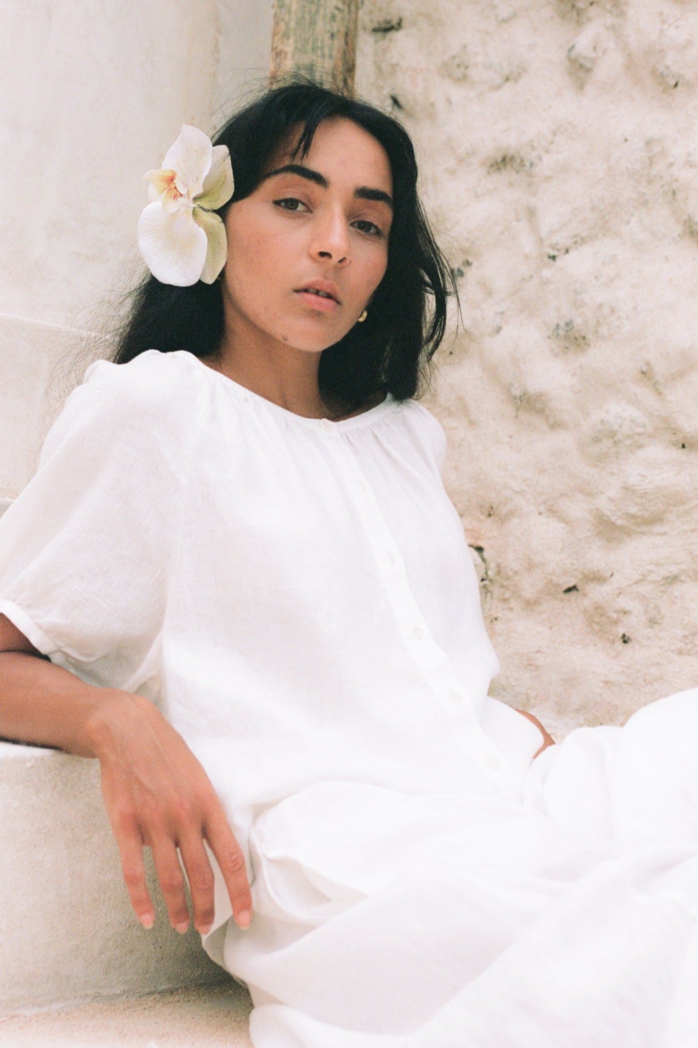 LILLY PILLY woman wearing an ivory Pia linen shirt top with a flower in her hair, sitting on steps by a textured wall